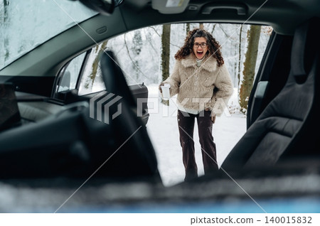 Woman with curly hair wearing a fluffy coat is standing outside a car in a snowy landscape, holding a cup while smiling during a winter day with trees in the background 140015832