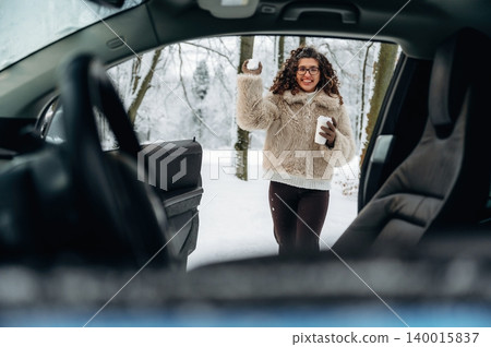 Smiling woman with curly hair wearing a cozy fur sweater is standing outside a car in a snowy landscape, holding a coffee cup and enjoying the winter scenery Smiling woman with curly hair wearing a cozy fur sweater is standing outside a car in a snowy landscape, holding a coffee cup and enjoying the winter scenery 140015837