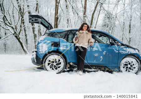 Electric car charging in snowy landscape with a woman wearing a fur coat standing beside the vehicle, showcasing winter conditions and eco-friendly transportation 140015841