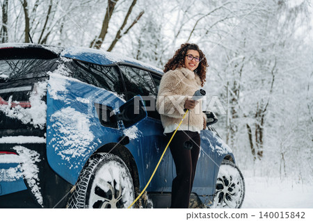 Woman with curly hair wearing a fur coat stands beside a blue electric car in a snowy landscape, holding a charging cable during winter weather with trees in the background 140015842
