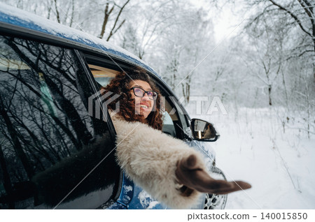 Young woman with curly hair wearing a warm coat is smiling and reaching out of a car window in a snowy landscape surrounded by trees during winter Young woman with curly hair wearing a warm coat is smiling and reaching out of a car window in a snowy landscape surrounded by trees during winter 140015850