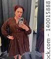 Woman takes a mirror selfie while wearing a brown embroidered dress in a fitting room. Fashion shopping, garment fitting, personal style, retail choice, and everyday wardrobe decision. 140015888