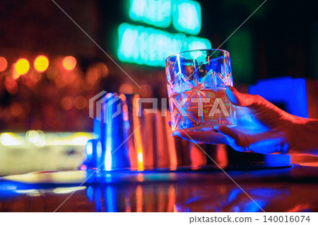 Hand taking whiskey glass from neon bar counter with colorful nightlife lights Hand taking whiskey glass from neon bar counter with colorful nightlife lights 140016074