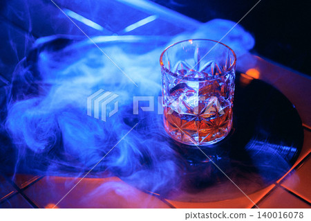 Blue smoke whiskey glass on vinyl record with dark neon nightlife mood. Blue smoke whiskey glass on vinyl record with dark neon nightlife mood. 140016078
