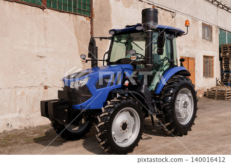 Blue Agricultural Tractor Parked Near Industrial Building in Sunny Daylight Blue Agricultural Tractor Parked Near Industrial Building in Sunny Daylight 140016412