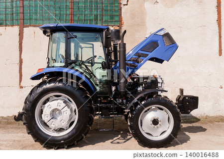 Blue Compact Tractor with Open Engine Hood Parked Near Rustic Building in Sunny Outdoor Setting Blue Compact Tractor with Open Engine Hood Parked Near Rustic Building in Sunny Outdoor Setting 140016488