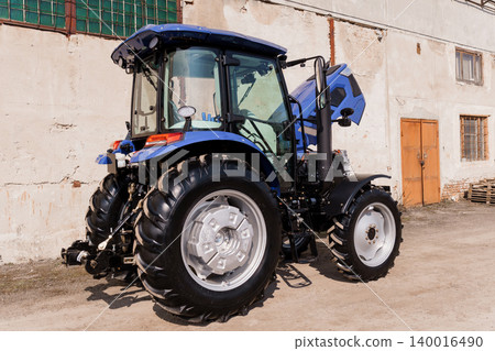 Blue Modern Agricultural Tractor Parked Outside Old Rural Farm Building in Sunlight Blue Modern Agricultural Tractor Parked Outside Old Rural Farm Building in Sunlight 140016490