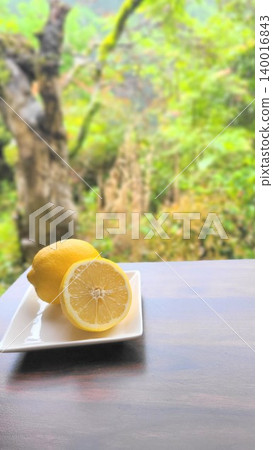 A vibrant lemon (whole and sliced) on a white plate against a backdrop of fresh greenery. 140016843