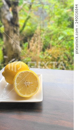 Against a backdrop of fresh greenery, vibrant lemons (whole and sliced) are arranged on a white plate. Against a backdrop of fresh greenery, vibrant lemons (whole and sliced) are arranged on a white plate. 140016844