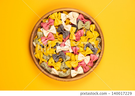 Multicolored farfalle pasta in wooden bowl on yellow background Multicolored farfalle pasta in wooden bowl on yellow background 140017229