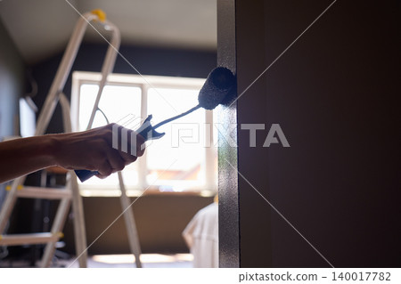 Hand uses paint roller to apply dark paint along wall corner, with ladder in background Hand uses paint roller to apply dark paint along wall corner, with ladder in background 140017782