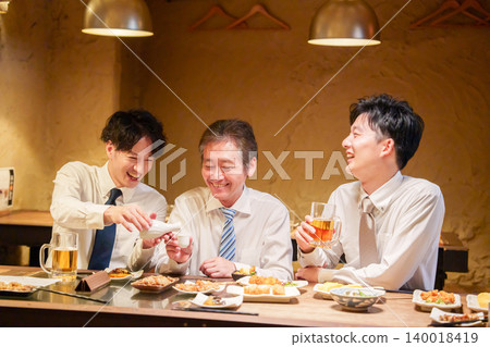 一位年輕員工在居酒屋(日本酒吧)為資深同事倒酒。 一位年輕員工在居酒屋(日本酒吧)為資深同事倒酒。 140018419