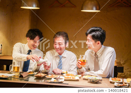 一位年輕員工在居酒屋(日本酒吧)為資深同事倒酒。 一位年輕員工在居酒屋(日本酒吧)為資深同事倒酒。 140018421