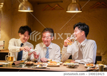 一位年輕員工在居酒屋(日本酒吧)為資深同事倒酒。 一位年輕員工在居酒屋(日本酒吧)為資深同事倒酒。 140018423
