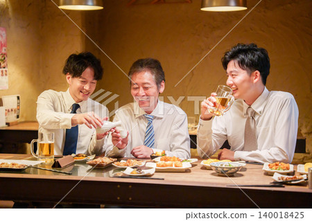 一位年輕員工在居酒屋(日本酒吧)為資深同事倒酒。 一位年輕員工在居酒屋(日本酒吧)為資深同事倒酒。 140018425