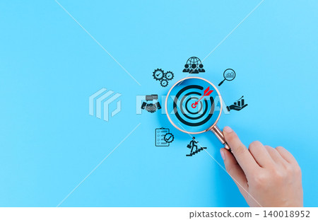 Business goals and target achievement concept. A hand holding a magnifying glass focuses on a target with an arrow and business icons like growth charts, gears, people, goal setting, and success. Business goals and target achievement concept. A hand holding a magnifying glass focuses on a target with an arrow and business icons like growth charts, gears, people, goal setting, and success. 140018952