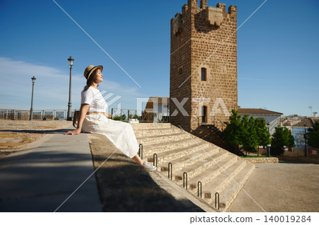 Woman traveling in a small Spanish medieval village sitting by a stone tower enjoying the view 140019284