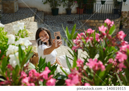 Woman traveling in a small Spanish village taking a selfie among flowers on a cobblestone street Woman traveling in a small Spanish village taking a selfie among flowers on a cobblestone street 140019315