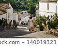 Woman walking down a cobblestone street in a small Spanish village with hat and basket 140019323