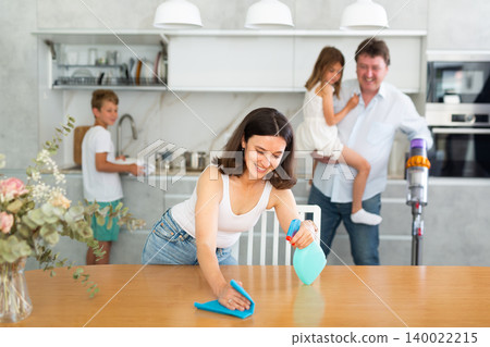 Wife cleans table with rag, husband and children clean kitchen in background Wife cleans table with rag, husband and children clean kitchen in background 140022215