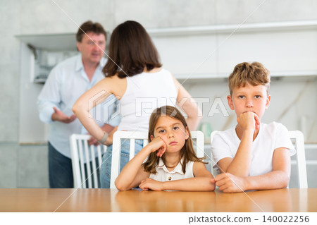 Frightened and frowning children sit at kitchen table during mom and dad quarrel. Frightened and frowning children sit at kitchen table during mom and dad quarrel. 140022256