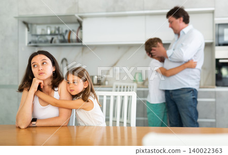 Offended mother sitting at the kitchen table being hugged by little daughter Offended mother sitting at the kitchen table being hugged by little daughter 140022363