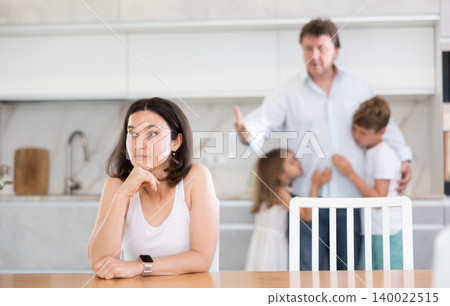 Offended wife sits at table and listens as her husband yells at her Offended wife sits at table and listens as her husband yells at her 140022515