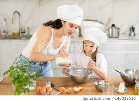 Happy mother and her daughter adding flour in metal bowl and whisking ingredients in the kitchen 140022516