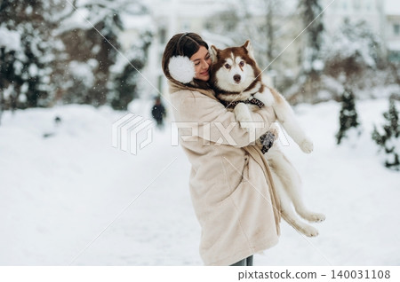 Woman with dark hair wearing a cream coat and earmuffs is holding a fluffy Siberian Husky in a snowy park with evergreen trees and soft snowfall in the background 140031108