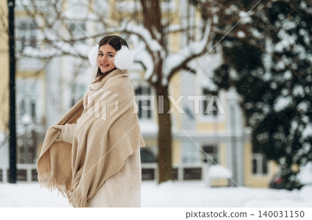 Young girl with dark hair wearing a warm beige shawl and earmuffs is smiling in a snowy outdoor setting with trees and buildings in the background during winter Young girl with dark hair wearing a warm beige shawl and earmuffs is smiling in a snowy outdoor setting with trees and buildings in the background during winter 140031150