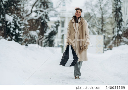 Young woman wearing winter clothing and earmuffs is walking through a snowy path carrying shopping bags in a winter landscape with trees and buildings in the background 140031169
