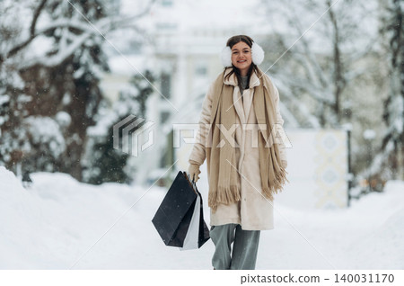Young woman with brown hair wearing winter clothing and earmuffs is walking through a snowy street carrying shopping bags during a winter day in a festive atmosphere Young woman with brown hair wearing winter clothing and earmuffs is walking through a snowy street carrying shopping bags during a winter day in a festive atmosphere 140031170