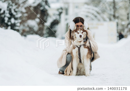 Young woman with dark hair wearing earmuffs is crouching beside a Siberian Husky dog on a snowy path surrounded by trees during winter weather Young woman with dark hair wearing earmuffs is crouching beside a Siberian Husky dog on a snowy path surrounded by trees during winter weather 140031178