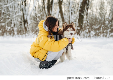Young woman in a yellow puffer jacket crouches in snow with a Siberian Husky dog, surrounded by a winter forest landscape during a sunny day Young woman in a yellow puffer jacket crouches in snow with a Siberian Husky dog, surrounded by a winter forest landscape during a sunny day 140031298