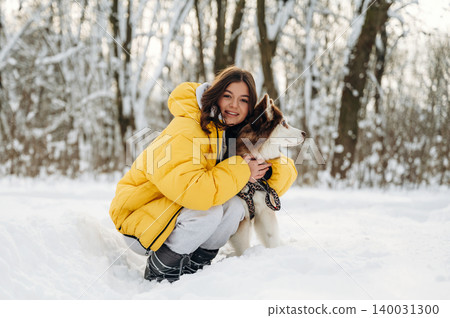 Young woman in a yellow puffer jacket is kneeling in the snow with a Siberian Husky dog, surrounded by trees in a winter landscape during a sunny day Young woman in a yellow puffer jacket is kneeling in the snow with a Siberian Husky dog, surrounded by trees in a winter landscape during a sunny day 140031300