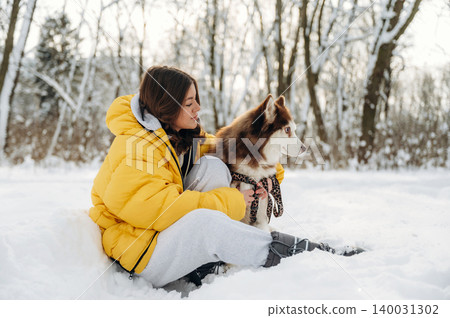 Young woman in a bright yellow puffer jacket sits in the snow with a Siberian Husky dog, surrounded by trees and a winter landscape during a cold day Young woman in a bright yellow puffer jacket sits in the snow with a Siberian Husky dog, surrounded by trees and a winter landscape during a cold day 140031302