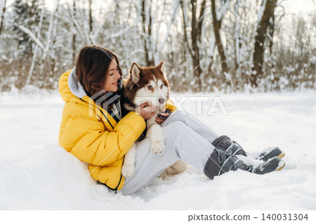 Young woman in a yellow puffer jacket sits in snow with a Siberian Husky dog, surrounded by trees and winter scenery, enjoying a playful moment outdoors Young woman in a yellow puffer jacket sits in snow with a Siberian Husky dog, surrounded by trees and winter scenery, enjoying a playful moment outdoors 140031304