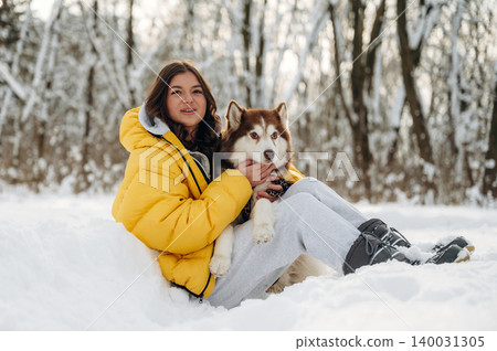 Young girl wearing a yellow puffer jacket is sitting in the snow with a Siberian Husky dog, surrounded by a winter forest landscape with snow-covered trees Young girl wearing a yellow puffer jacket is sitting in the snow with a Siberian Husky dog, surrounded by a winter forest landscape with snow-covered trees 140031305