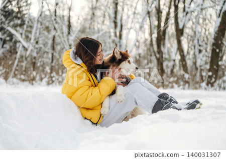Young girl wearing a yellow puffer jacket sits in the snow with a brown and white dog, surrounded by trees covered in snow during a winter day Young girl wearing a yellow puffer jacket sits in the snow with a brown and white dog, surrounded by trees covered in snow during a winter day 140031307