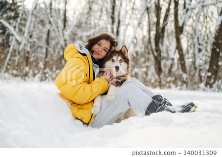 Young woman with brown hair wearing a yellow puffer jacket is sitting in the snow with a Siberian Husky dog during a winter day in a snowy forest setting Young woman with brown hair wearing a yellow puffer jacket is sitting in the snow with a Siberian Husky dog during a winter day in a snowy forest setting 140031309