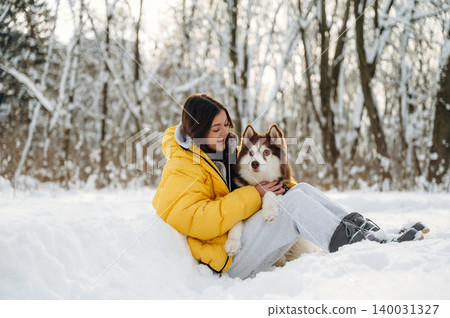 Young woman in a bright yellow puffer jacket sits in the snow with a brown and white husky dog, surrounded by a winter landscape of trees and snow-covered ground Young woman in a bright yellow puffer jacket sits in the snow with a brown and white husky dog, surrounded by a winter landscape of trees and snow-covered ground 140031327