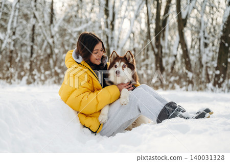 Young woman with dark hair wearing a yellow puffer jacket is sitting in the snow with a brown and white husky dog during a winter day in a snowy forest Young woman with dark hair wearing a yellow puffer jacket is sitting in the snow with a brown and white husky dog during a winter day in a snowy forest 140031328