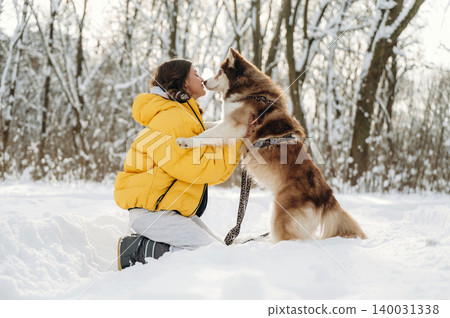 Young girl wearing a yellow puffer jacket kneels in the snow while playing with a brown and white Siberian Husky dog in a snowy forest setting with trees in the background Young girl wearing a yellow puffer jacket kneels in the snow while playing with a brown and white Siberian Husky dog in a snowy forest setting with trees in the background 140031338