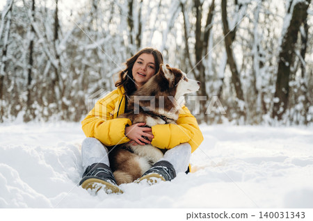 Young woman in a bright yellow jacket sits in the snow with a brown and white dog, surrounded by trees covered in snow during a winter day in a forest setting 140031343
