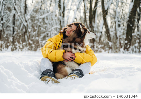 Young woman in a bright yellow jacket sits in the snow with a fluffy brown and white dog, surrounded by a winter forest landscape with snow-covered trees 140031344