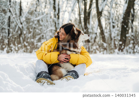 Young woman with long dark hair wearing a yellow puffer jacket is sitting in the snow while hugging a brown and white dog in a winter forest setting with trees in the background Young woman with long dark hair wearing a yellow puffer jacket is sitting in the snow while hugging a brown and white dog in a winter forest setting with trees in the background 140031345