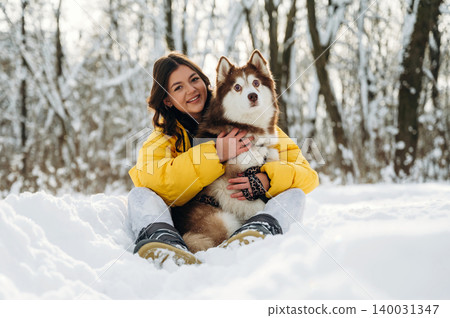 Young woman with dark hair wearing a yellow puffer jacket is sitting in the snow while holding a brown and white husky dog in a winter forest setting with snow-covered trees Young woman with dark hair wearing a yellow puffer jacket is sitting in the snow while holding a brown and white husky dog in a winter forest setting with snow-covered trees 140031347