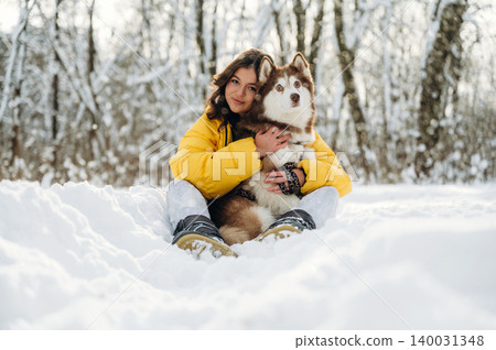 Young woman with long brown hair wearing a yellow jacket is sitting in the snow while holding a fluffy brown and white dog in a winter forest setting with trees in the background Young woman with long brown hair wearing a yellow jacket is sitting in the snow while holding a fluffy brown and white dog in a winter forest setting with trees in the background 140031348