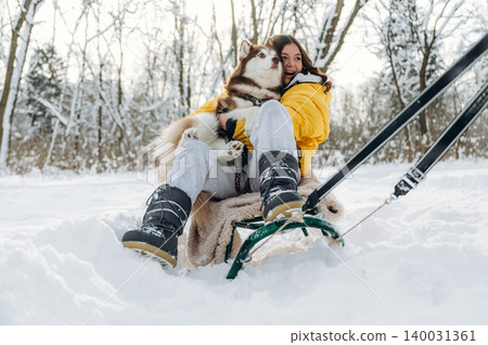 Young woman in yellow winter jacket sits on a sled in the snow with a fluffy dog, surrounded by trees and a winter landscape during a sunny day 140031361