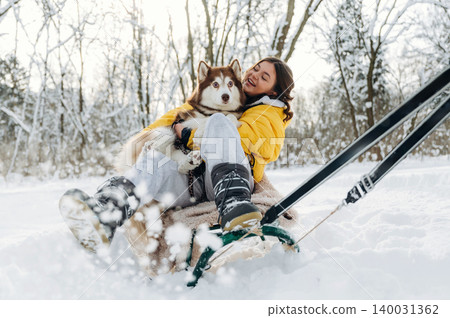 Young woman in yellow jacket sitting on a sled with a Siberian Husky dog in a snowy landscape surrounded by trees during a winter day with soft sunlight 140031362